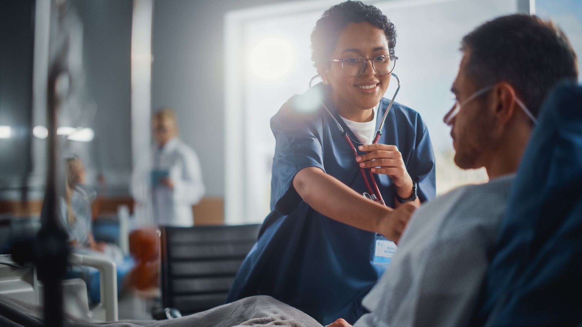 Hospital Ward: Friendly Black Head Nurse Uses Stethoscope to Listen to Heartbeat and Lungs of Recovering Male Patient Resting in Bed, Does Checkup. Man Getting well after Successful Surgery